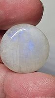 Close-up photo of a round, polished rainbow moonstone gemstone showing blue flash and internal inclusions held between fingers.