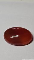 Oval red carnelian cabochon on white surface, showing smooth polished face with rich red-orange color.