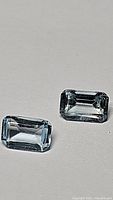 Two emerald cut sky blue topaz gemstones on a neutral background, displaying clarity and cut.