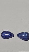 Two pear-shaped cabochon tanzanite stones shown together on neutral background. Purple-blue color with translucency and inclusions visible.