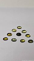 Ten oval cabochon cut tourmaline gemstones arranged in a triangular pattern on a white surface, showing their deep yellow to green coloring with some visible inclusions.