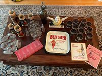 Overview of all barware items on wooden table including steins, glasses, bottle, novelty stein, tray, and pamphlets