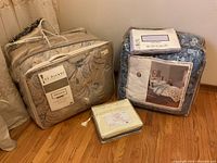 Two comforter sets packaged, one beige paisley Stonebriar and one blue floral Vintage Bliss Toscana, plus a packaged set of sheets in front on wood floor.