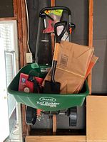 Front view of green Scotts Turf Builder EdgeGuard DLX spreader with shovel, sprayer, and paper bags inside against a wooden wall.