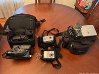 Photo showing all cameras and bags on a wooden table, including a Hitachi camcorder and two digital cameras, with multiple black carrying cases.