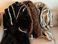 Three women's faux fur coats arranged on a couch showing varied prints and colors, including black and white, leopard print, and light tan fur