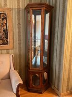 Full front view of the wooden curio cabinet with glass sides, situated next to a chair and patterned wallpaper.