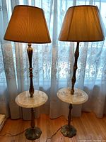 Pair of vintage floor lamps with fabric shades, metal bases, and built-in round marble tables. Lamps standing side by side near window with lace curtains.