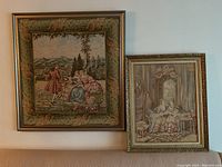 Two framed needlepoint or tapestry artworks hanging on a wall showing classical figures in historic dress, larger piece on left, smaller on right