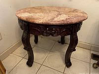 Front-angle view of round wooden table with marble top showing carved legs and apron with floral motifs.