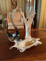 Lot on wooden table showing three Murano glass pieces: a black swan figurine, a white/orange marbled vase, and a clear decorative bowl.