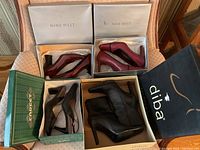 Four pairs of women's shoes in boxes on a chair: two pairs of red high heel pumps by Nine West, one pair of black ankle boots by Diba, one pair of taupe and black pumps by Funatics.