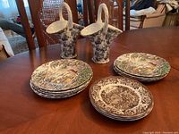 Two ceramic pitchers with twisted handles and black and white floral pattern, surrounded by multiple decorative plates with countryside and village scenes in brown and multicolor.