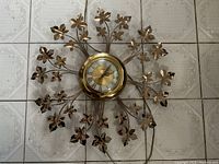 Full frontal image of the United wall clock lying on a patterned tile floor showing the entire clock face and metal leaf decorations woven on stems extending radially.