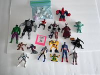 Photo displaying a variety of small action figures including Hulk, Rhino-armored figure, Spiderman, small white monster figures in bag, and other figures like Bumblebee robot.