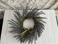 Overhead view of artificial lavender wreath with green leaves and olive green satin bow against white background and neutral fabric underneath.