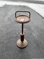 Front view of ashtray stand showing the full height, circular ridged base, central support column with a central band detail, round tray top with a handle under natural lighting outdoors.