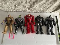 Five large Marvel Avengers action figures displayed in standing position on white board. Figures include Thanos, Hulk, Iron Man Hulkbuster, and two War Machine.