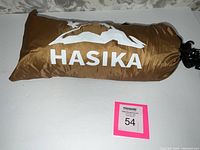 Brown nylon carry bag with Hasika logo containing the camping tent. Bag shows a drawstring closure with a black hook attached for hanging or securing.