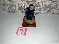 Photo of small ceramic sculpture titled "Rebocito" by Todo Padilla. Figure dressed in blue holding a white cloth with signature plate on wooden base.
