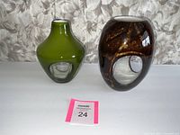 Two art glass vases, one green with smooth glossy finish and hollow clear base, one brown with swirling pattern and clear base with hollow detail.