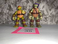 Two Teenage Mutant Ninja Turtle figures, Raphael and Donatello, standing on a white surface with a MaxSold tag in front.