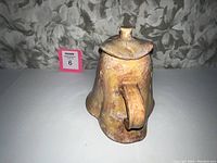 Front view of unique studio pottery pitcher with handle and lid, showing earthy glaze and irregular shape.