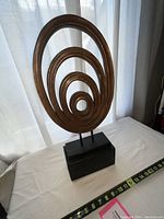 Wood art sculpture showing concentric oval rings arranged from smallest to largest on a black rectangular base, with a tape measure for size reference.