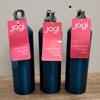 Three Jogi branded dark blue stainless steel water bottles with rings on screw caps and attached pink product tags.