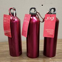 Three identical magenta Jogi stainless steel water bottles standing upright. Each has a ring top black screw cap with carabiner and product tags attached.
