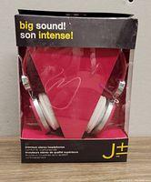 Front view of the headphones inside clear plastic packaging, showing the white cushioned ear pads, red triangular background, and black outer box with yellow and white text.