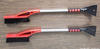 Two new 32-inch snow brushes with black bristles, red plastic handle accents, aluminum shafts, and black plastic ice scrapers on opposite ends, placed on wood flooring.