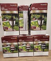 Photo showing six boxed Paradise brand low voltage 12V landscape lighting fixtures from the Estate Series, various styles including post lanterns, modern path light, and round zone lights in cast aluminum with UV protected finish.