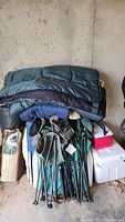 Photo showing stacked camping chairs, sleeping bags on top, cooler, rolled tent or tarp in carrying case, and a red container box in a storage space with concrete walls and floor.