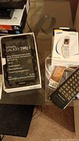 Photo shows a Samsung Galaxy Tab 2 sealed packaging, Samsung flip phone with user guide, and several remote controls on glass table.