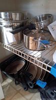 Overhead shelf view showing stainless steel cookware including saucepans, double boiler, and frying pans stacked on a wire rack shelf.