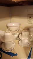 Stack of white ceramic dishes including plates and bowls, cups and saucers stacked nearby.