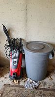 Pressure washer and Rubbermaid Brute garbage can side by side next to wall on dirty floor with some yard debris