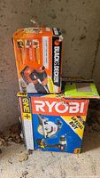 Photo of Ryobi OnePlus 18V drill and circular saw kit box stacked with Black & Decker finishing sander box and sandpaper pack on concrete floor with dirt and leaves around.