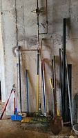 Photo showing various garden tools including rakes, hoes, shovels, metal poles leaning against a concrete wall.
