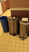 Photo showing two stainless steel trash cans with foot pedals and one tall blue plastic trash can in a kitchen corner on tiled floor.