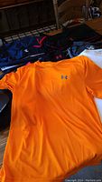 Men's Under Armour athletic clothing including short sleeve shirts in orange, white, red, navy, pants, and accessories laid out on a wooden table and nearby surfaces.