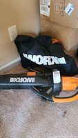 Worx blower with black carrying bag showing the WORX logo in white letters. Unit has black and orange details, placed on carpet with some items in background.