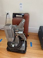 Full view of Revere P-90 8mm projector with brown carrying case behind it on wood floor.