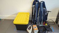 Photo of the lot against a wall, showing two folded blue easy-open cots with carry bags, two yellow-lid black storage boxes, a bike pump, gloves, flashlight, small knapsack, and bags with bungee cords and emergency supplies.