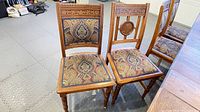Group of wooden dining chairs with carved backs and patterned seats arranged in a line, showing various carvings and upholstery patterns.