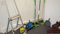 Overall lot showing the small aluminum step ladder, various mops, brooms, dustpans, and cleaning supplies lined up against a wall.