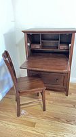 Secretary desk with the writing surface folded down, showing interior drawers and shelving, paired with matching wooden chair.