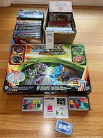 Photo showing DVDs, VHS tapes, Beyblade Burst QuadDrive toy set, two tray puzzles, and electronic word game on a wooden surface.