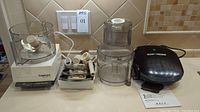 Cuisinart food processor base with attached processor bowl on left and extra processor bowl on right along with blades and accessories, and George Foreman grill unit.